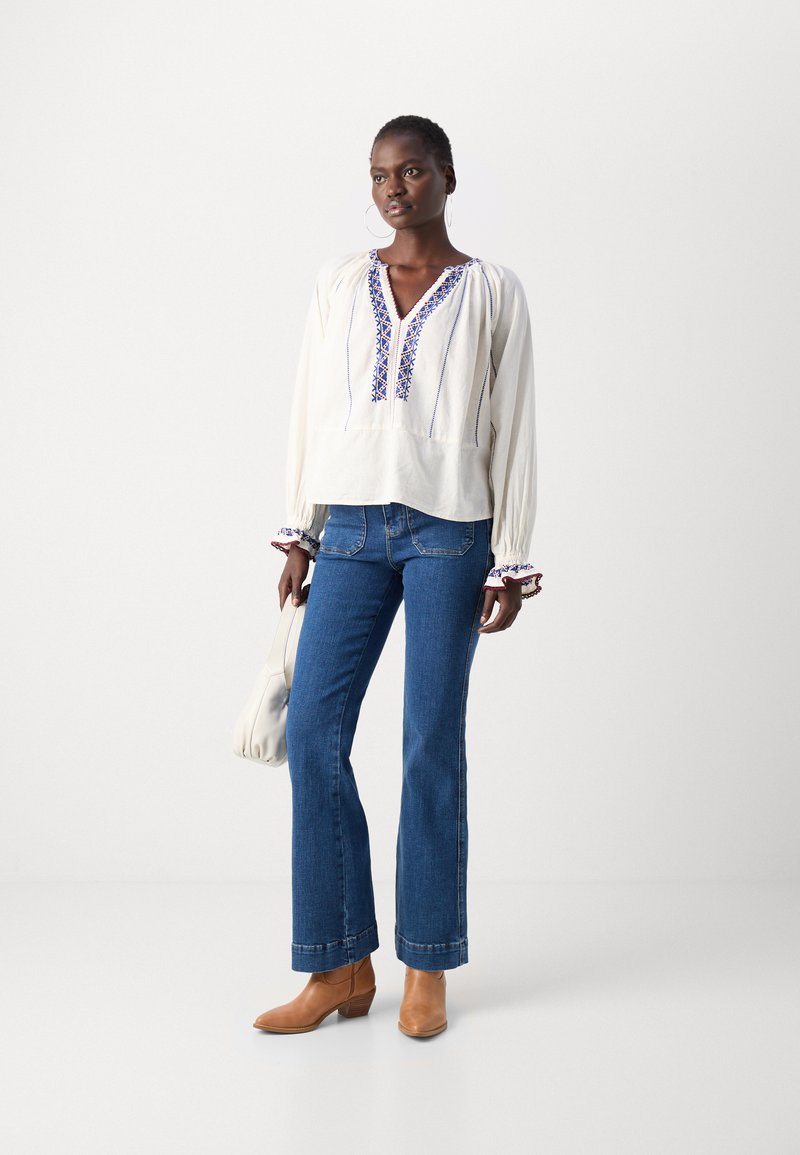 Blouse blanche à manches longues avec broderies bleues, associée à un jean bleu évasé et des bottines brunes. La blouse a des manches froncées et un col en V.