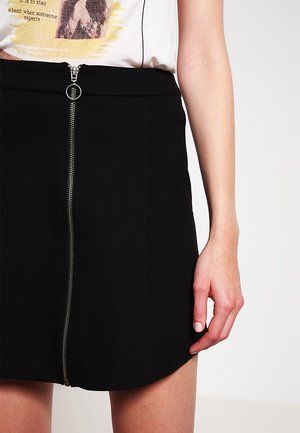 Femme portant une jupe noire taille haute avec une fermeture éclair argentée à l'avant et un t-shirt blanc à motif.
