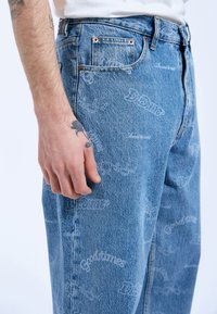 Jeans de mezclilla azul claro con un patrón de texto blanco en toda la prenda. Incluye bolsillos tradicionales y detalles metálicos.