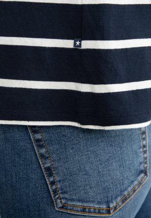 Tumed ja valged triibulised särk, mille allservas on väike tähte logo, koos siniste teksadega, millel on klassikaline tagatasku disain.