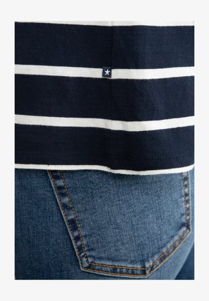 Jūras zila un balta svītrainā krekls ar mazu zvaigznes logo apakšējo malā, apvienots ar zilu džinsu biksēm ar klasisku aizmugurējā kabatas dizainu.