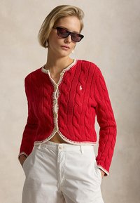 Kobieta w czarnych okularach przeciwsłonecznych, czerwonym skróconym swetrze na drutach z białą lamówką oraz białych spodniach z wysokim stanem, stojąca z rękami w kieszeniach.