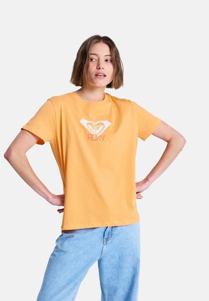 T-shirt en coton jaune à manches courtes avec un imprimé cœur blanc et logo. Le modèle porte un jean bleu clair, mains sur les hanches, sur fond blanc.