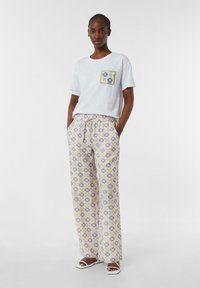 Biała koszulka z wzorzystą kieszenią, w połączeniu z luźnymi, wzorzystymi spodniami w lawendowym i żółtym kolorze; modelka nosi białe sandały.