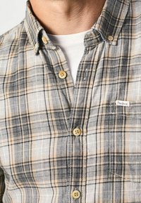 Camicia a quadri in grigio, nero e beige con colletto button-down, maniche corte e taschino sul petto. Presenta bottoni color sabbia e tessuto testurizzato.