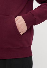 Sudadera burdeos con un bolsillo frontal, puños acanalados y dobladillo. La tela parece suave, con una textura lisa y un detalle de costura duradero.
