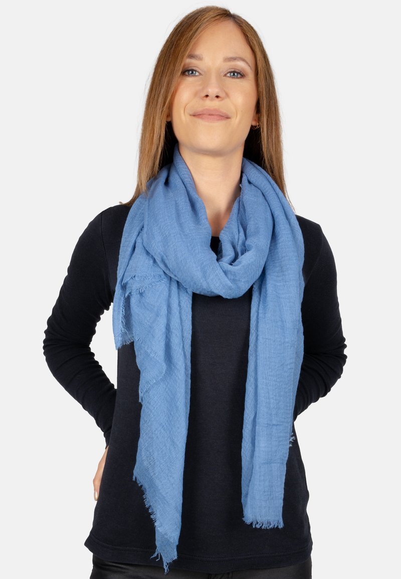 Écharpe bleu clair en tissu doux et texturé, avec des bords effilochés et un tomber détendu, portée de manière lâche autour du cou.