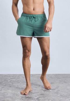 Skjorteløs mand stående barfodet på gråt gulv iført grønne badeshorts med hvid kant, hænderne i lommerne, mod en lysegrå baggrund.