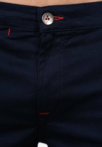 Pantaloni blu navy con dettagli di cucitura rossa e un bottone metallico in vita. Il tessuto è liscio con un design sartoriale.
