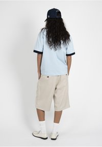 Personne aux longs cheveux bouclés portant une casquette bleu marine à l’envers, une chemise bleu clair, un short beige jusqu’aux genoux, des chaussettes blanches et des chaussures beiges, vue de dos.