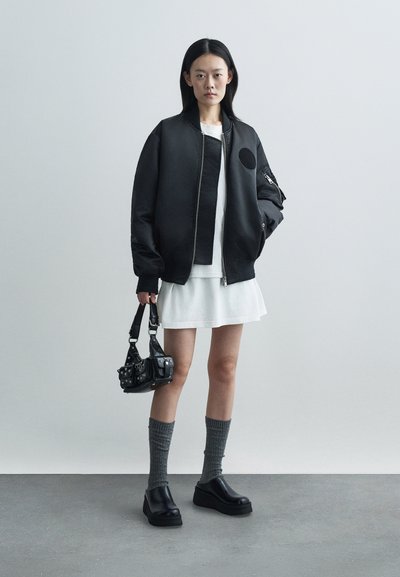 Črn bombnik s okroglo nalepko, kombiniran z belo obleko, sivimi golfnimi nogavicami do kolen in robustnimi črnimi platform čevlji. Mala črna torbica.