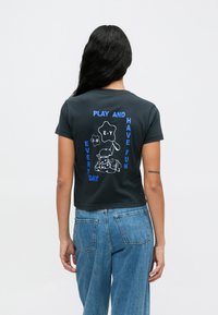 Svart kortermet t-skjorte med hvite og blå tegneserieillustrasjoner, teksten lyder "LEK OG HA MORO HVER DAG," med lekne karakterdesigns.