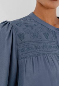 Blaue Bluse mit gestickten Blumenmustern auf der Schulter und der Brust, Rundhalsausschnitt und gerafften Puffärmeln.