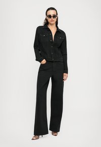 Woman wearing black denim jacket and wide-leg jeans with open-toe heels and small black sunglasses, posing against plain white background.