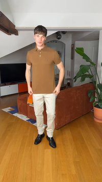 Mladi moški, oblečen v rjavo polo majico, bež hlače in črne čevlje, stoji v sodobni dnevni sobi z rastlino in oranžnim kavčem.