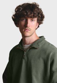 Sudadera cremallera verde oliva con cuello; presenta una cremallera de metal y tejido texturizado; se lleva encima de una camiseta de color claro.