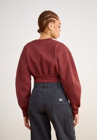 Bordeaux színű cropped kapucnis pulóver széles ujjal és elasztikus aljjal, szürke cargo nadrággal párosítva, amelyen egy zseb található és diszkrét textúrával rendelkezik.