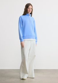 Γυναίκα που στέκεται φορώντας ανοιχτό μπλε φούτερ με φερμουάρ και άσπρο παντελόνι με φαρδιά πόδια, μπροστά σε λευκό φόντο.