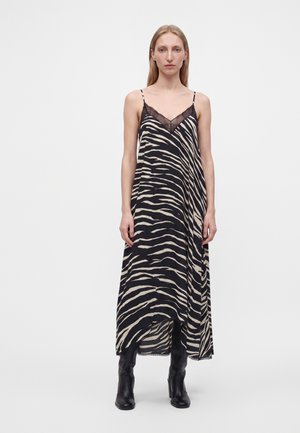 Femme debout portant une robe sans manches rayée zèbre noire et beige avec bordure en dentelle, associée à des bottes noires montant jusqu’aux genoux.