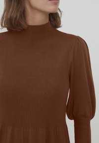 Bruine geribde jurk met hoge hals, lange pofmouwen en een uitlopende rok. Zachte textuur met een aansluitend lijfje en geplooide mouwdetails.