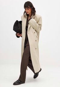Beige double-breasted coat with gold buttons, paired with a brown suit and black flat shoes; holding a black clutch bag.