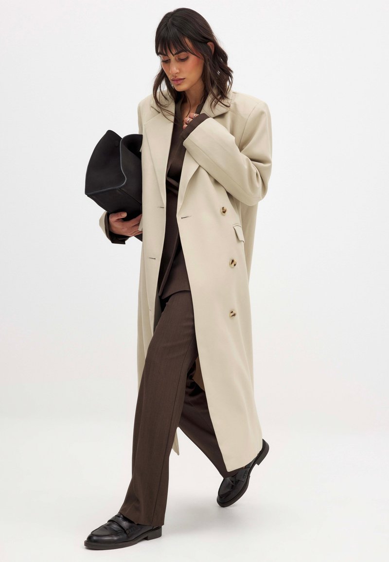 Beige double-breasted coat with gold buttons, paired with a brown suit and black flat shoes; holding a black clutch bag.