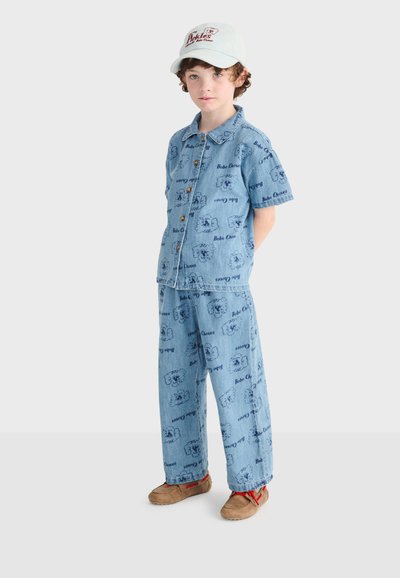 Enfant portant une chemise et un pantalon en denim bleu clair avec un imprimé répétitif de dessins animés, des chaussures beiges et une casquette blanche, debout les mains derrière le dos.