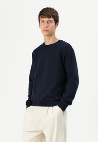 Sweatshirt bleu marine en tissu doux, avec un col rond et des poignets et un ourlet côtelés, porté avec un pantalon beige clair.