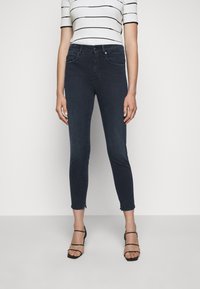 Jeans ajustados de mezclilla oscura con dobladillo deshilachado, combinados con una blusa blanca de rayas. Presenta cinco bolsillos y un cierre frontal con botón.
