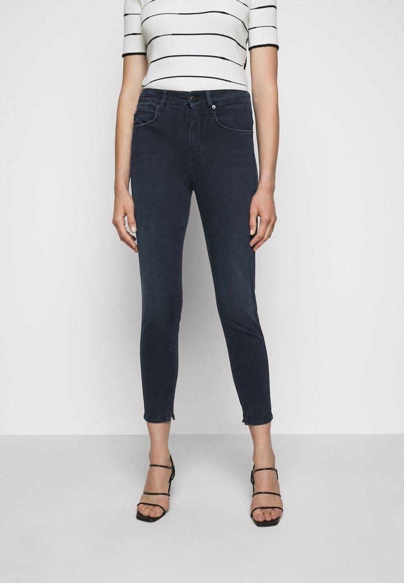 Jeans ajustados de mezclilla oscura con dobladillo deshilachado, combinados con una blusa blanca de rayas. Presenta cinco bolsillos y un cierre frontal con botón.