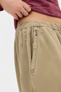 Pantaloni beige a coste con vita elasticizzata e tasche laterali, dotati di un piccolo cartellino con logo sul davanti. Tessuto morbido e texturizzato.