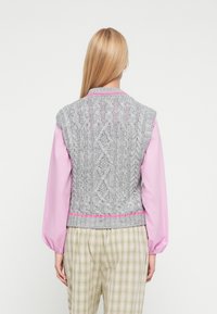 Szary dziergany bezrękawnik z warkoczowym wzorem, okrągłym dekoltem i bez rękawów. Posiada różowy akcent wzdłuż brzegu. Noszony na różowej koszuli.