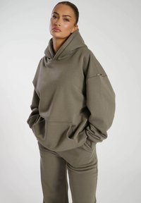 Sudadera de gran tamaño en color verde oliva, hecha de una tela suave, con un bolsillo frontal, mangas anchas y un gran escote en forma de capucha. Combinada con pantalones a juego.