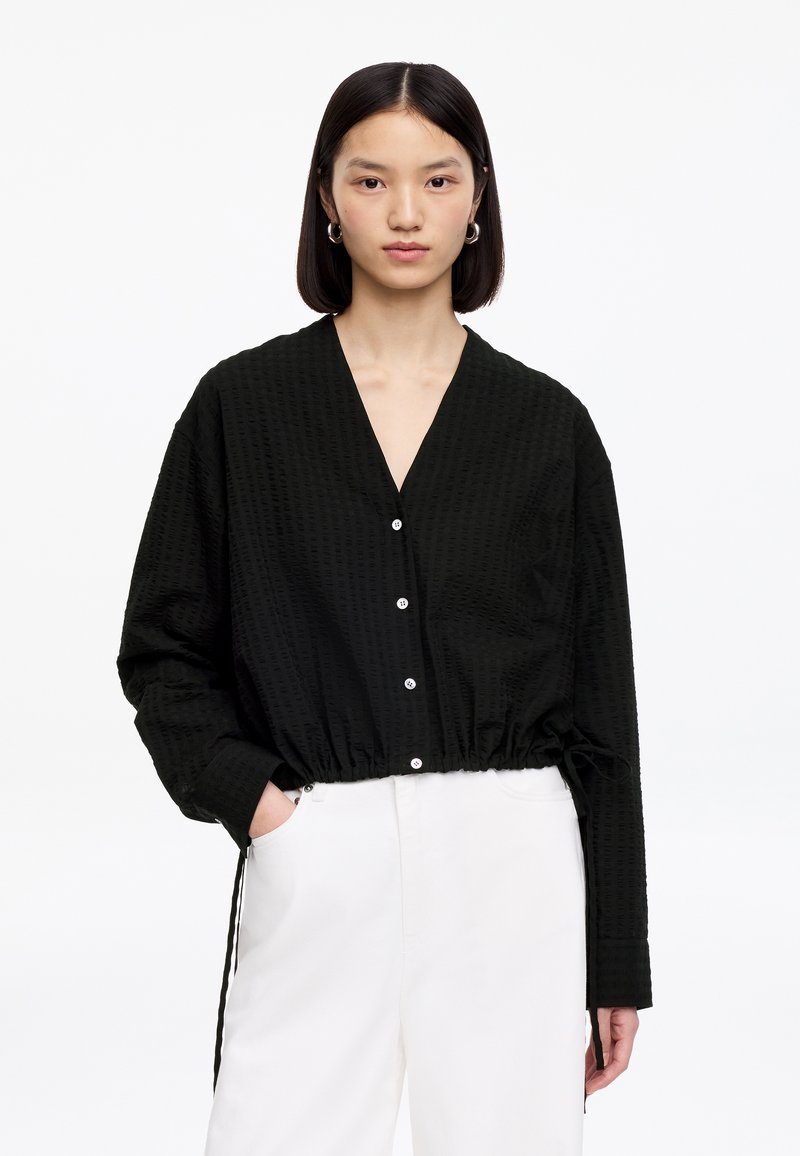 Jeune femme aux cheveux noirs lisses portant un haut noir ample à boutons et un pantalon blanc, debout devant un fond blanc uni.