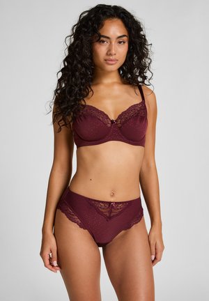 Vrouw met lang krullend haar, gekleed in een bijpassende bordeauxrode kanten lingerie set, staand tegen een effen grijze achtergrond, met het gezicht naar voren gericht.