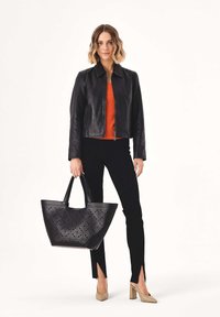 Frau trägt schwarze Lederjacke, orangefarbenes Oberteil, schwarze Hosen mit vorderen Schlitzen, beige Absätze und hält große schwarze Tragetasche mit ausgeschnittenem Muster.