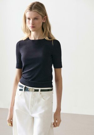 Femme aux longs cheveux blonds portant un haut noir à manches courtes ajusté, un pantalon blanc taille haute et une ceinture noire à boucle argentée.