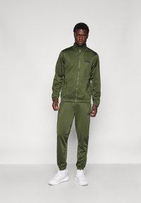 Olijfgroen trainingspak van gladde stof; ritsjack en elastische broek met geribde boorden, gecombineerd met witte sneakers.