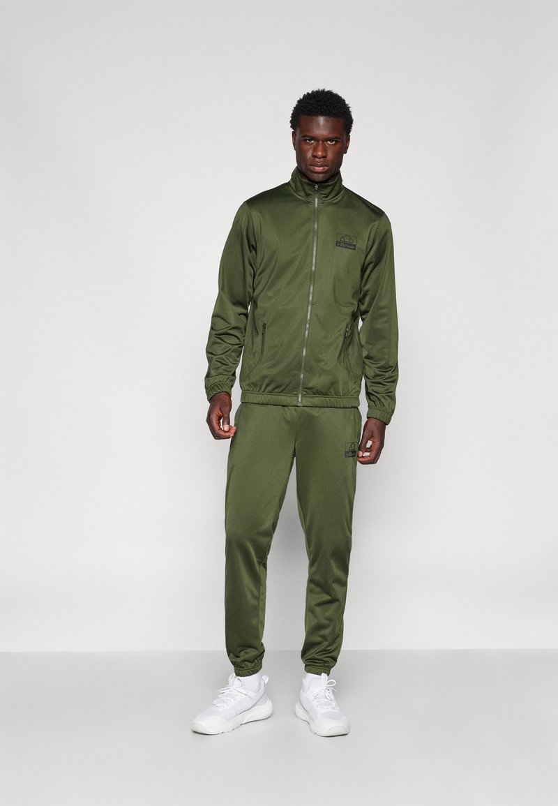Olijfgroen trainingspak van gladde stof; ritsjack en elastische broek met geribde boorden, gecombineerd met witte sneakers.