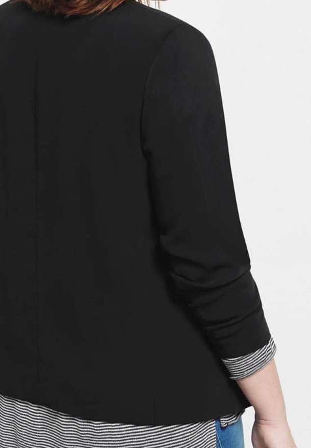 Personne portant un blazer noir sur une chemise rayée noir et blanc, vue de dos avec le bras gauche légèrement plié.
