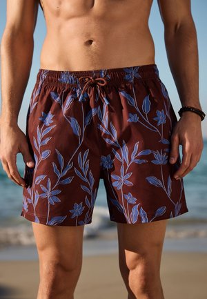 Primo piano di un uomo che indossa pantaloncini da bagno color vinaccia con motivi floreali blu, in piedi su una spiaggia con onde oceaniche sullo sfondo.