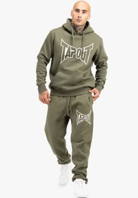 Olivengrønt hoodie- og sweatpants-sæt med hvid "TAPOUT" logo. Lavet af blødt stof, med en frontlomme og snøre i taljen. Hvide sneakers.