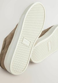 Chaussures en daim marron avec semelles en caoutchouc blanc affichant un motif de bande de roulement triangulaire et la marque "NEXT" au centre de la semelle.