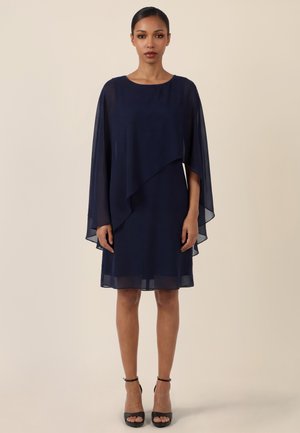 Frau trägt ein Kleid mit marineblauem Chiffonüberzug und asymmetrischem Saum sowie schwarze Knöchelriemen-Pumps, steht vor einem schlichten beigen Hintergrund.