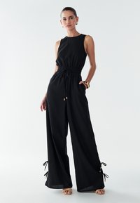 Mono negro sin mangas con cintura elástica y cordón. Piernas anchas con lazos ajustables en los puños. Tejido liso con un ajuste relajado.