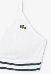 Bralette cropped blanco con textura acanalada, tirantes ajustables, logo de cocodrilo verde en la copa izquierda y banda acanalada con franjas verdes y blancas.