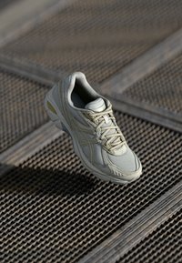 Ljusbeige sportskor med en texturerad mesh-överdela, vadderad tunga och snören, med subtila gula detaljer på sulan.