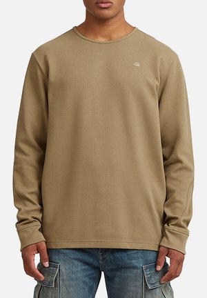 Homme portant un t-shirt à manches longues en maille gaufrée beige et un jean cargo bleu, debout les bras détendus le long du corps.