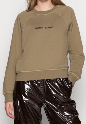 Femme portant un sweat-shirt beige avec le texte « SAMSOE SAMSOE » et un pantalon en vinyle marron foncé brillant, une main dans la poche, torse visible.