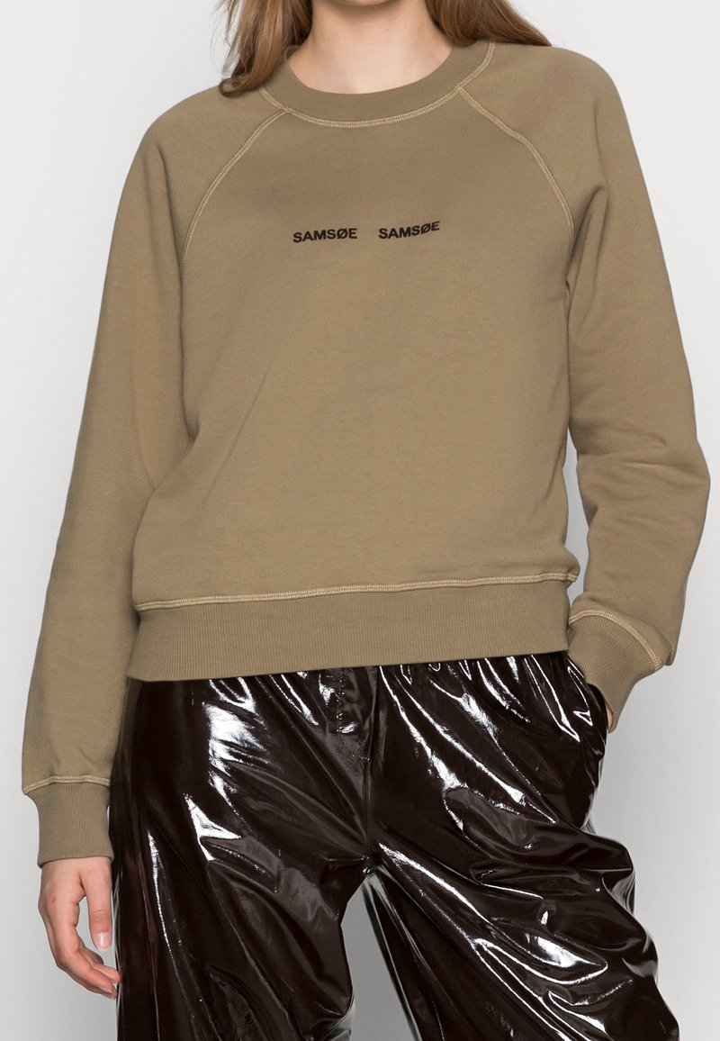 Femme portant un sweat-shirt beige avec le texte « SAMSOE SAMSOE » et un pantalon en vinyle marron foncé brillant, une main dans la poche, torse visible.